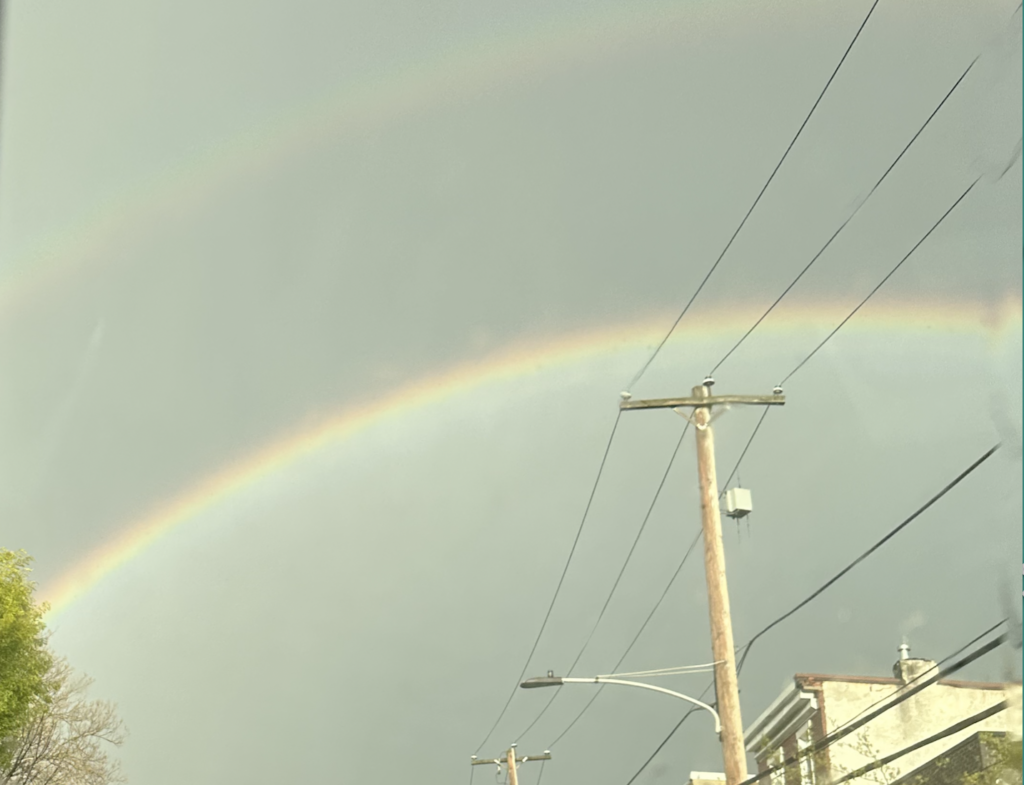 A double rainbow appears in a cloudy sky above utility poles and buildings.